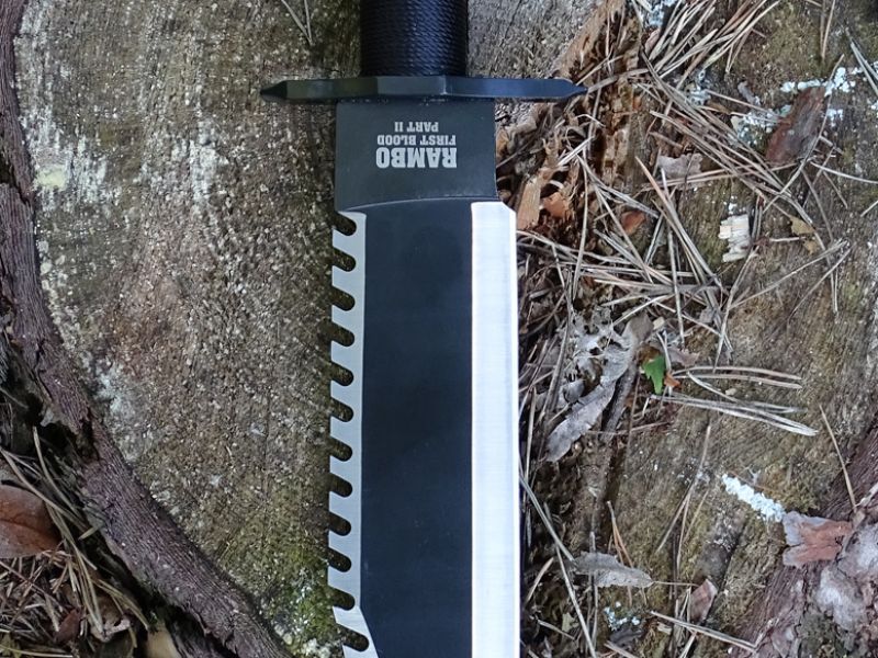 Dvojí boční pohled na nůž RAMBO FIRST BLOOD PART II. Jednak bez pouzdra a jednak zasunut ve svém černém pouzdře. I u dvojky je replika pouzdra vyrobena z umělé kůže. I zde ve slušné kvalitě, ale obdobně jako u jedničky i zde chybí kapsa na brousek.