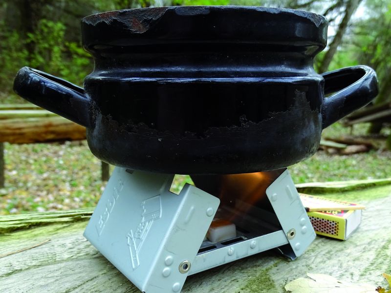 Pohled na malý hrnec o objemu cca 0,5 litrů vody na malém vařiči v ne úplném otevření. Malý vařič je optimální pro jednu až dvě osoby. Dutinu obou vařičů můžete využít pro uskladnění jednak samotného tuhého lihu, sirek, čaje, cukru apodobně. V báglu je nutno využít každičkej kousek volného místa.